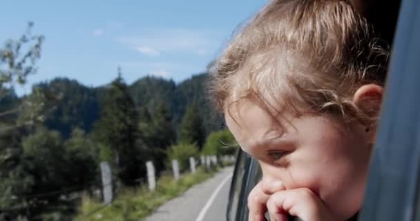 Glad söt liten flicka med ett leende som sticker ut genom bilfönstret mot vinden. Långsamma rörelser V2 — Stockvideo