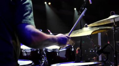 Canlı konser performansı, dramatik ışıklandırma altında enerjik bir şekilde sahnede çalan bir davulcu. Sahne, davullara ve zillere vuran bagetlerin dinamik hareketini yakalıyor. Yüksek kalite 4k görüntü