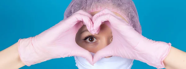 Close-up of the beautiful positive face of a doctor or nurse, hands in pink nitrile gloves holding the shape of a heart next to the eye on the blue banner panorama Healthy heart. Mock up