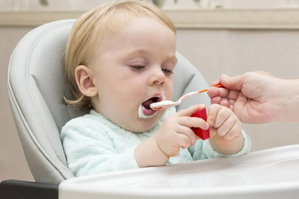 A mother feeds her son in a special baby feeding chair with a small spoon. The mother gives the child food with a child\'s spoon. Happy baby eats with an appetite.