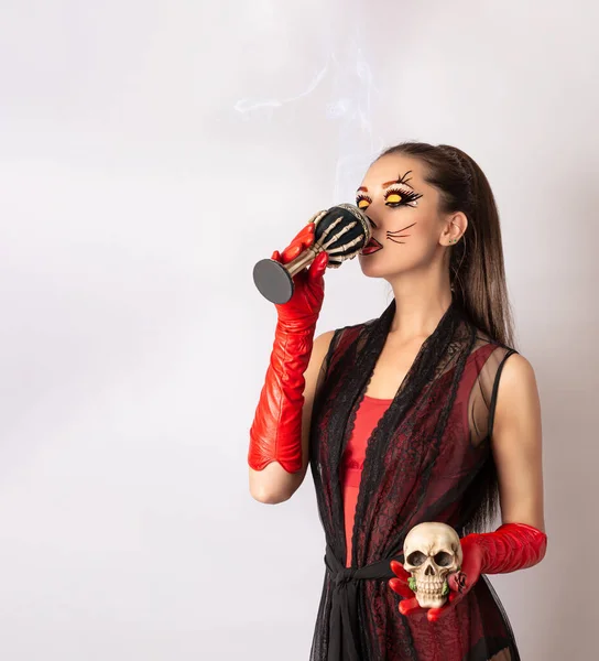 A girl with a painted face in a black dress and red gloves, a Cup of steaming drink in her hand, and a skull in the other hand. Face painting for Halloween. Costume for a masquerade.