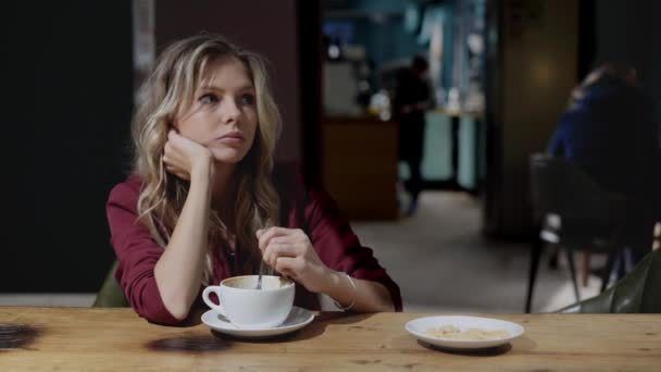 Грустная молодая женщина с вьющимися волосами пьет кофе в кафе и думает: — стоковое видео