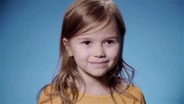 Милая маленькая девочка со светлыми волосами улыбается синий фон замедленной съемки — стоковое видео
