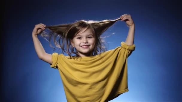 Χαριτωμένο αστείο μικρό κορίτσι παίζει με μακριά μαλλιά, μπλε φόντο αργή κίνηση — Αρχείο Βίντεο