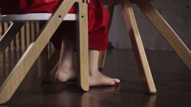 Menina bonito em uma saia vermelha sentado em uma mesa e acenando com os pés descalços — Vídeo de Stock