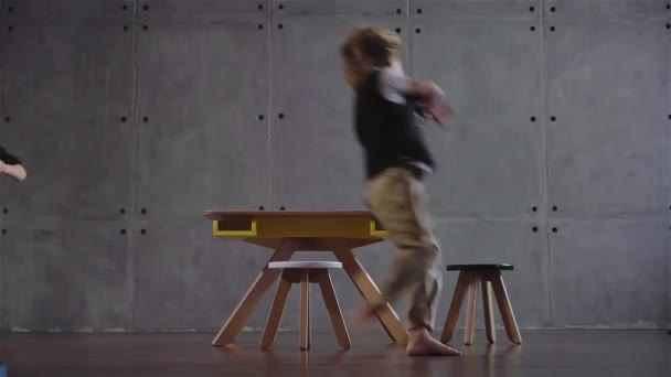 Dois meninos correndo em torno de uma mesa em uma sala de parede cinza — Vídeo de Stock