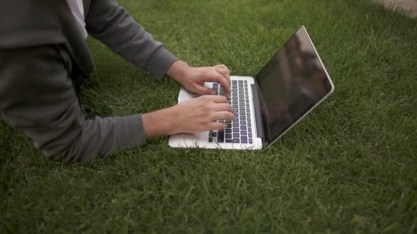 Невпізнаваний молодий чоловік, що друкує на ноутбуці, лежить на газоні, вид збоку — стокове відео
