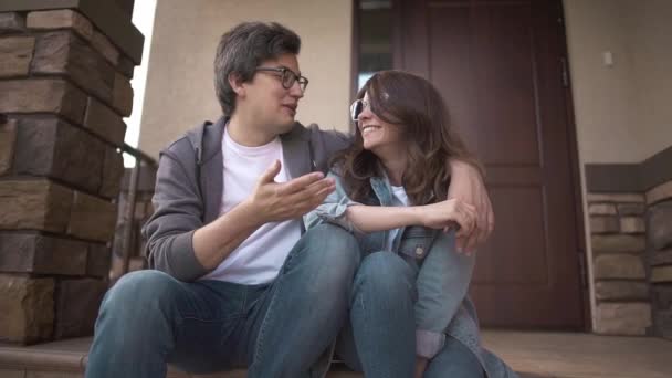 Skrattar unga paret sitter tillsammans på deras hus veranda och pratar — Stockvideo