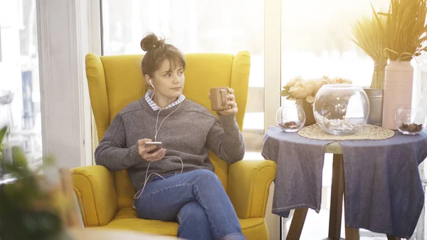 Çay içme müzik telefon ile büyük bir sandalyede oturan bir genç kız — Stok fotoğraf