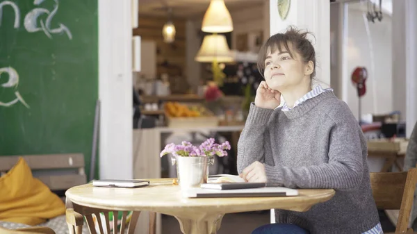 Una giovane ragazza bruna carina sorridente è seduta in un caffè a riflettere — Foto Stock