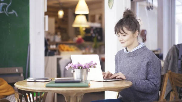 Una joven morena linda sonriente está trabajando con un ordenador portátil en un café — Foto de Stock