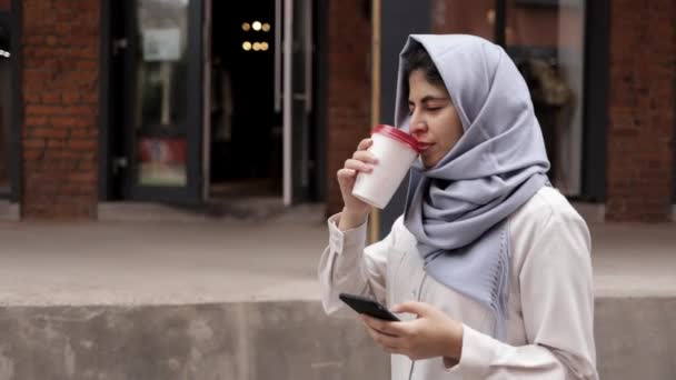 Jovem no hijab bebendo um café para ir e olhando para o smartphone — Vídeo de Stock