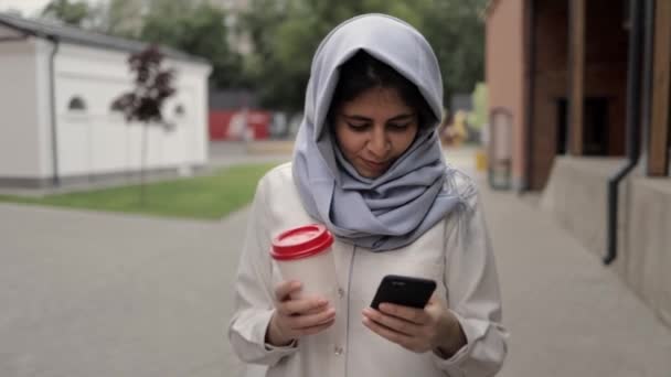 Mujer joven en hijab bebiendo café y mirando el teléfono celular — Vídeos de Stock