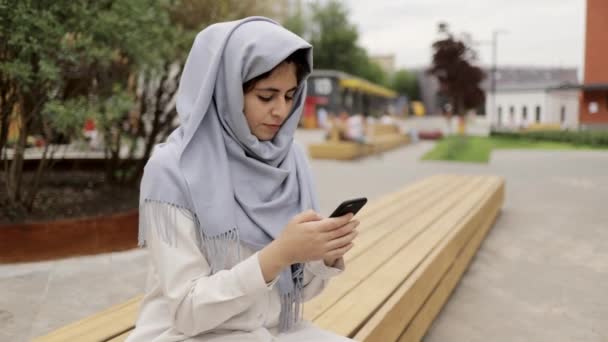 Молода жінка в сірому хіджабі смс на лавці в місті — стокове відео