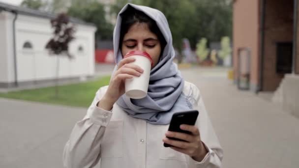 Mosolygó fiatal nő hidzsáb kávét iszik, és látszó-on sejt telefon — Stock videók
