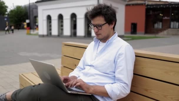 Jovem empresário de óculos digitando sentado em um banco na cidade — Vídeo de Stock