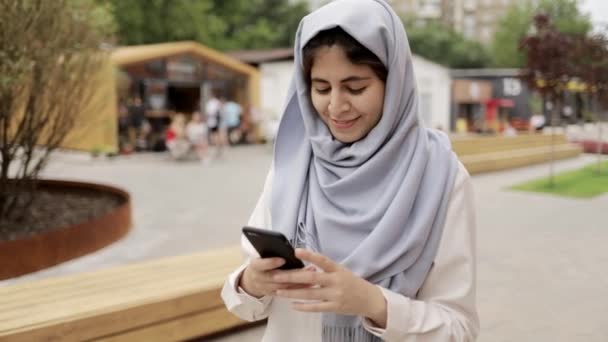 Glad ung kvinna i hijab kontrollera hennes smartphone i en sommar street — Stockvideo