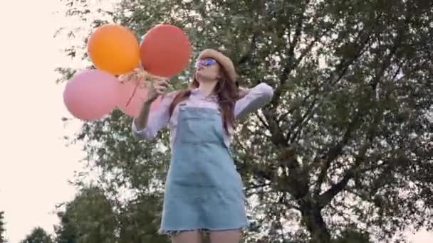 Улыбающаяся рыжая женщина в солнечных очках, стоящая с воздушными шарами в летнем парке — стоковое видео