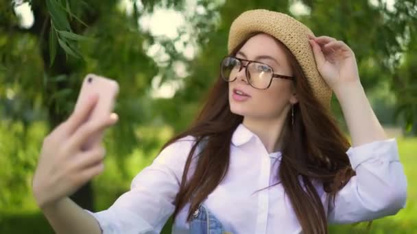 Рыжая девушка в очках и соломенной шляпе делает селфи в парке — стоковое видео