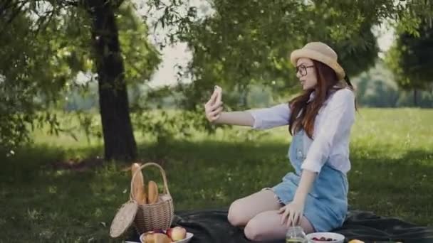 Chica pelirroja en gafas y sombrero de paja haciendo una selfie en el picnic en el parque — Vídeo de stock