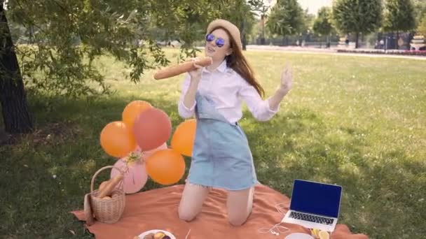 红头发的年轻女子戴着紫色的太阳镜 一顶草帽和一条牛仔裤裙 在夏日公园里用法式面包作为麦克风 唱歌跳舞 滑块中枪 — 图库视频影像