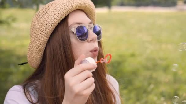 Hipster meisje met rood haar blazen van zeepbellen in zomer park — Stockvideo