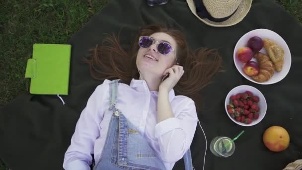 Chica joven de jengibre en gafas de sol y overoles tumbados en la hierba escuchando música — Vídeo de stock