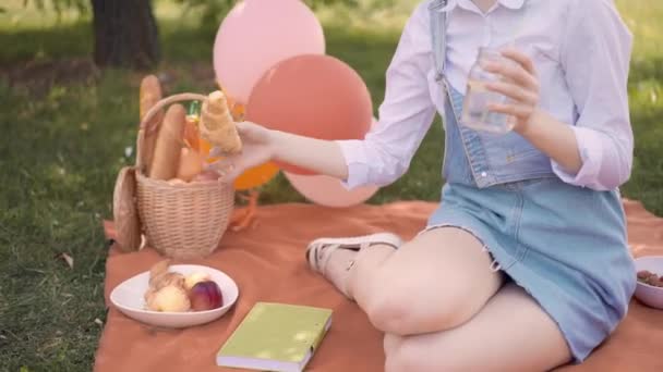 Рыжая девушка ест круассан и пьет сок в летнем парке, пикник — стоковое видео