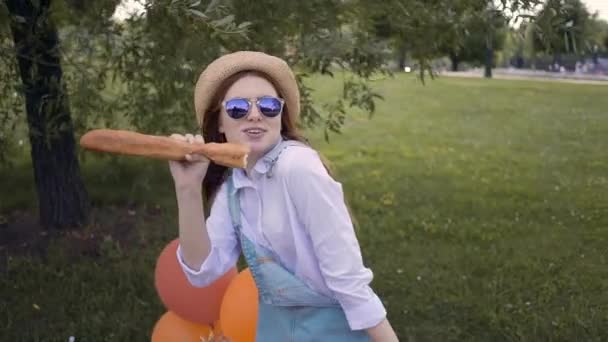 Hipster girl singing with a French bread as a mike in summer park, picnic — Stock Video