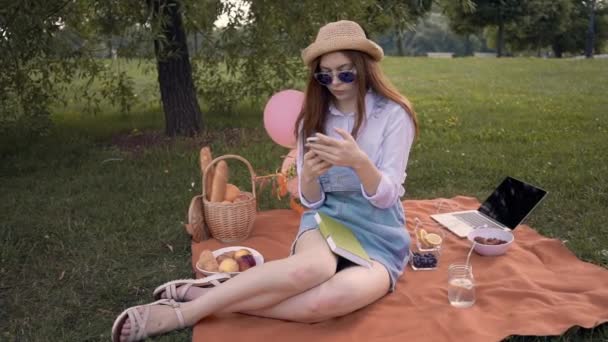 Ginger college student in straw hat and sunglasses texts and studies in park — Stock Video