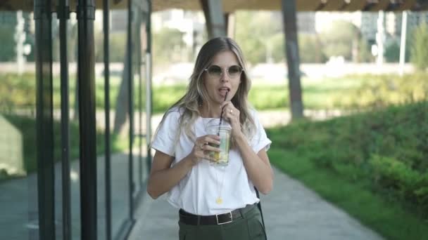 Hermosa mujer rubia en gafas de sol bebiendo bebida fría y caminando en el parque — Vídeos de Stock