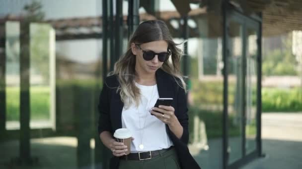 Mujer rubia en gafas de sol con café caminando y mirando el teléfono inteligente — Vídeos de Stock