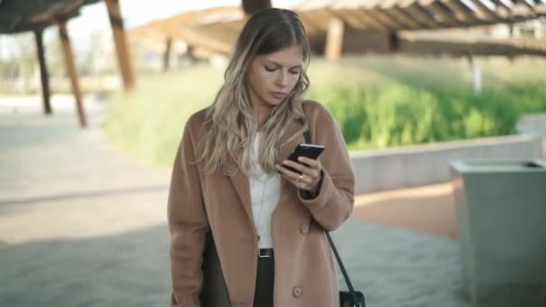 Привлекательная блондинка в пальто ходит и смотрит на свой смартфон осенью — стоковое видео