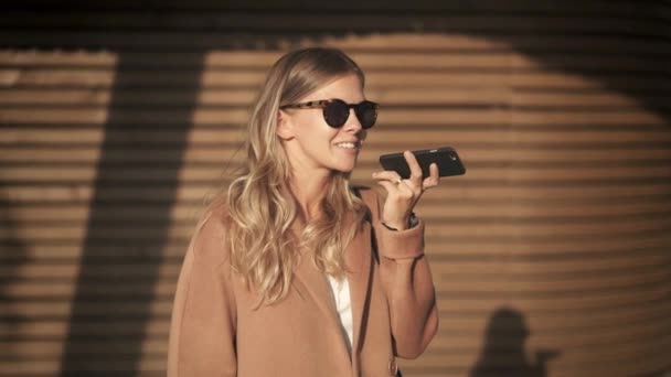 Блондинка в пальто и солнцезащитных очках разговаривает по телефону на осенней улице — стоковое видео