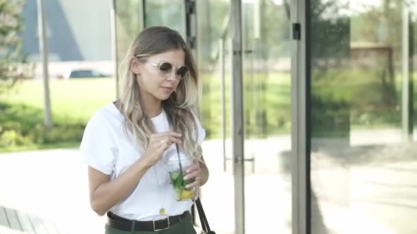 Hermosa mujer rubia en gafas de sol bebiendo mohito frío y caminando en el parque — Vídeos de Stock