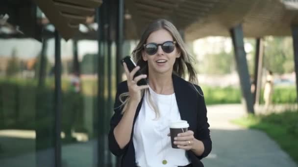 Blondine mit Sonnenbrille spaziert mit Kaffee und spricht auf Smartphone — Stockvideo