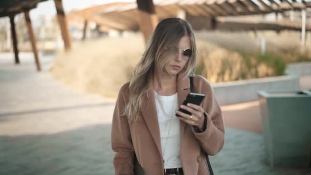 Mooie blonde vrouw met vacht lopen en kijken naar haar smartphone in de herfst — Stockvideo