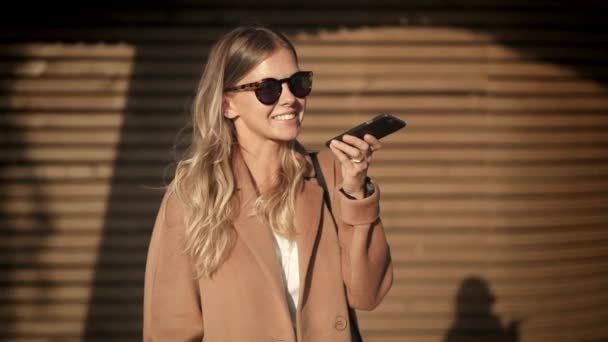 Chica rubia alegre en abrigo y gafas de sol hablando por teléfono en la calle de otoño — Vídeo de stock