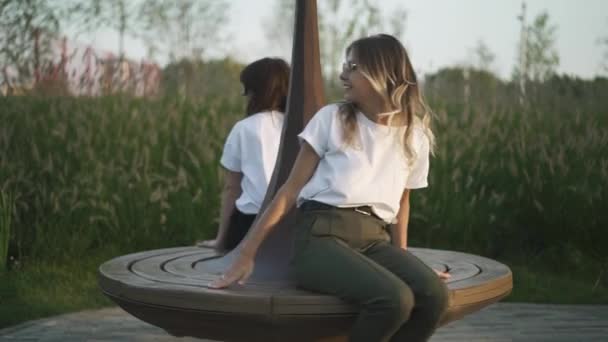 Две молодые девушки в футболках и солнцезащитных очках кружатся на скамейке в парке — стоковое видео