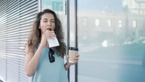 Красивая молодая женщина ест круассан и пьет кофе, проходя мимо здания — стоковое видео