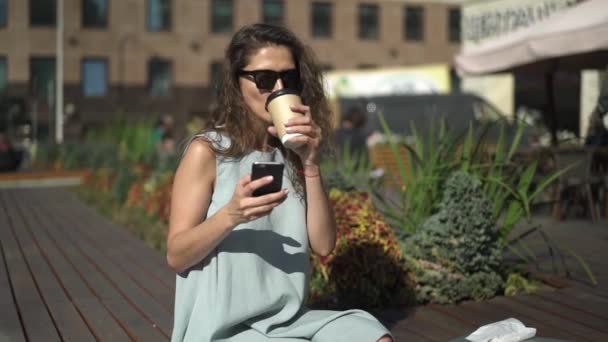 Hermosa joven bebiendo café y mirando el teléfono cerca de la cafetería al aire libre — Vídeos de Stock