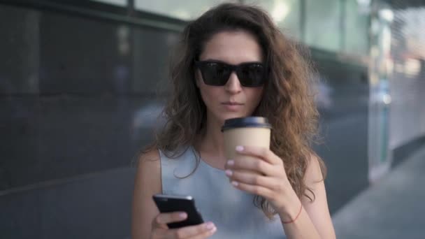 Ελκυστική νεαρή κοπέλα γραπτών μηνυμάτων και πίνοντας καφέ στέκεται στο δρόμο το καλοκαίρι — Αρχείο Βίντεο