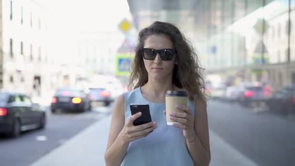 Mooie vrouw in zonnebril texting en drinken koffie wandelen in de zomer straat — Stockvideo