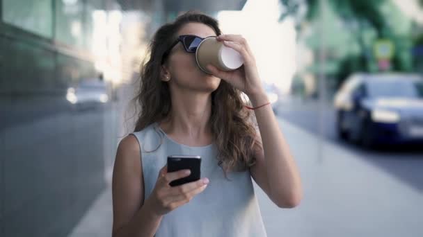 Mooie vrouw in zonnebril texting en drinken koffie staande in zomer straat — Stockvideo