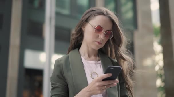 Mujer joven con gafas de sol rojas mirando su teléfono inteligente de pie en la calle de la ciudad — Vídeos de Stock