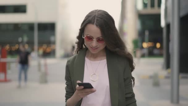 Mujer de negocios con teléfono caminando en la calle borrosa de la ciudad y sonriendo — Vídeos de Stock