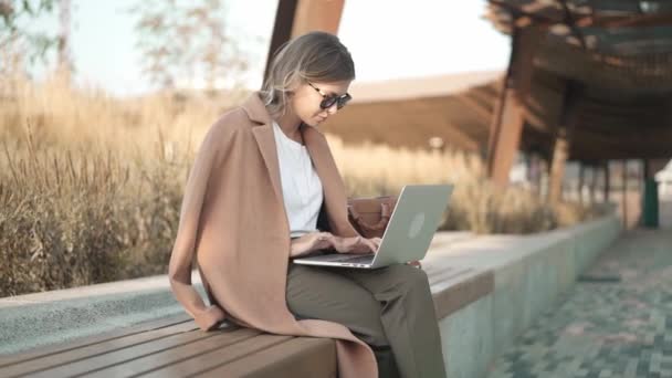 Mujer rubia calma en abrigo beige trabajando con el ordenador portátil en el parque — Vídeos de Stock