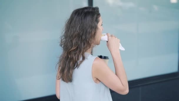 Young fair hair woman eating croissant and drinking coffee passing by building — Stock Video