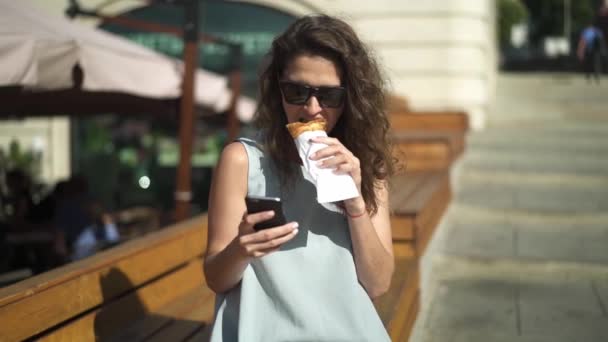 Mulher bonita comendo croissant e olhando para o telefone perto de café ao ar livre — Vídeo de Stock