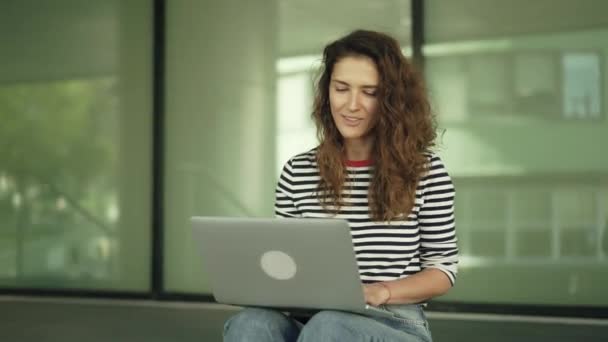 Wesoła, młoda kobieta w dżinsy wideokonferencji do jej przyjaciel siedzi poza — Wideo stockowe